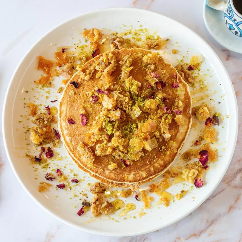 Mediterranean pancakes topped with crushed pistachios, walnuts, honey, and rose petals on a white plate.