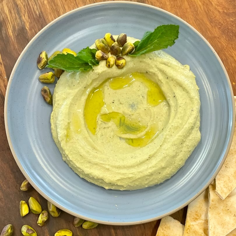 Pistachio hummus with olive oil, fresh mint, and pistachios on a blue plate with pita bread.