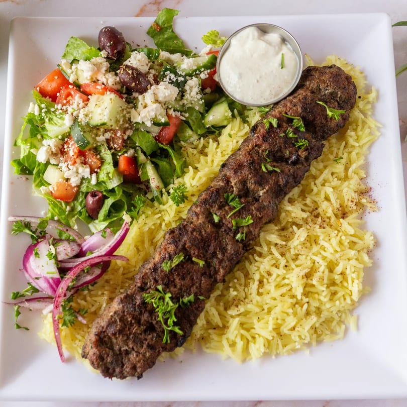 Mediterranean kofta kebab with rice, Greek salad, red onions, and a side of white sauce.