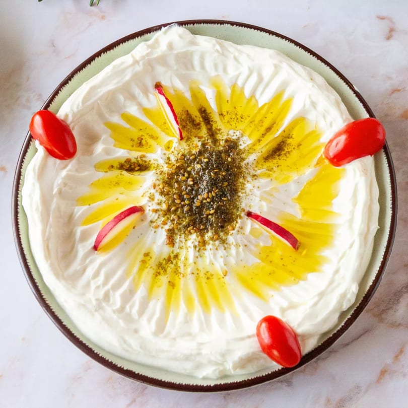 Creamy labneh dip with olive oil, za'atar, cherry tomatoes, and radish slices on a light green plate.