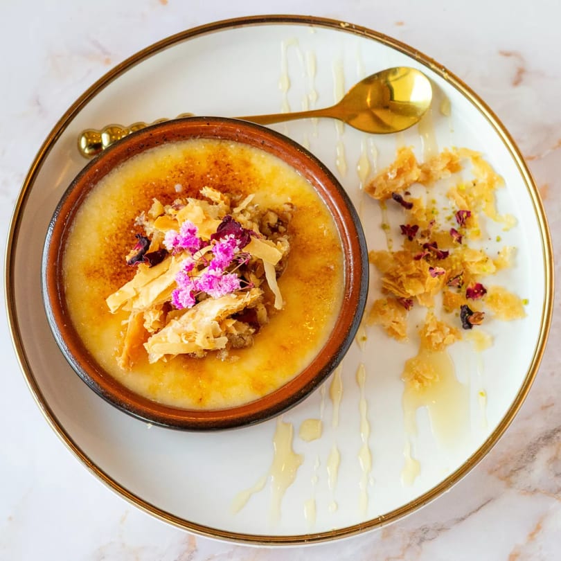 A Mediterranean dessert, likely creme brulee, topped with pink flowers and crispy pastry on a white plate.
