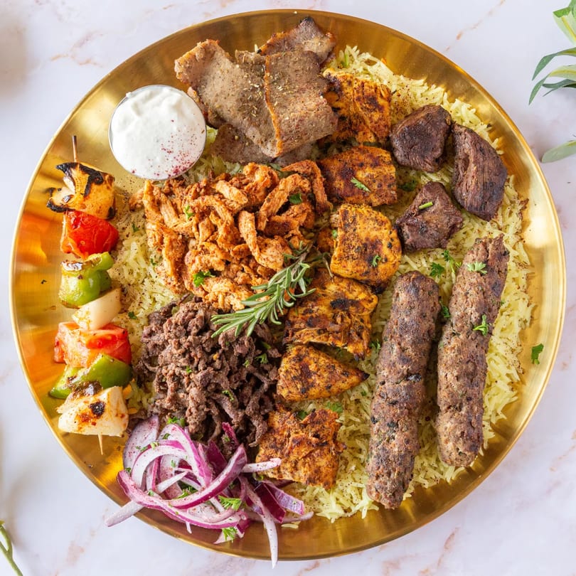 A large golden platter of Mediterranean grilled meats, rice, and vegetables, with a side of white sauce.